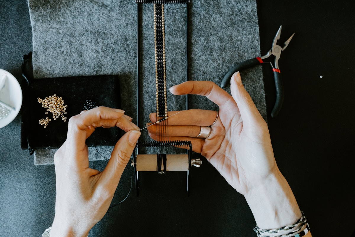 Mains manipulant un outil de couture, perles et accessoires sur fond gris.
