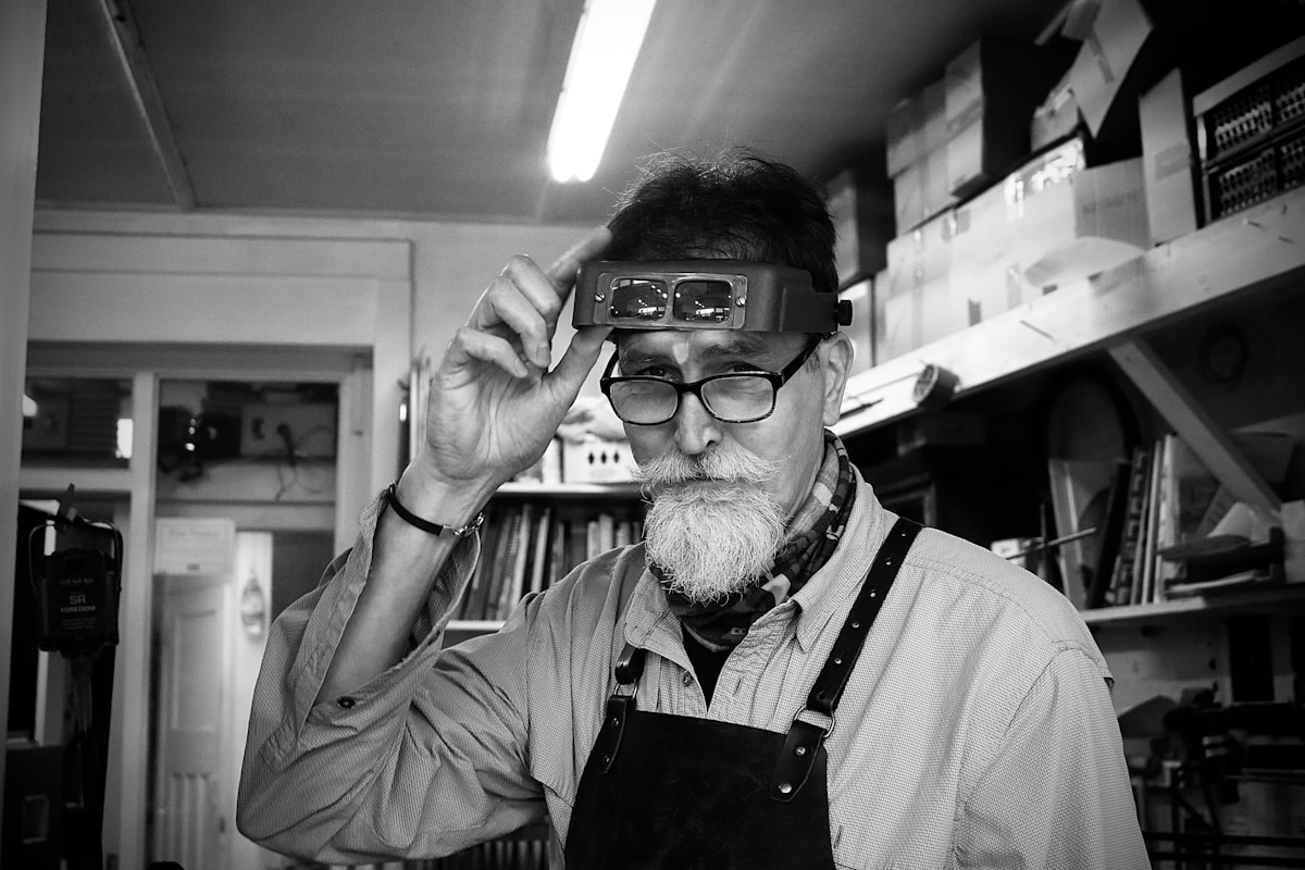 Homme âgé avec barbe, lunettes et loupe, dans un atelier.
