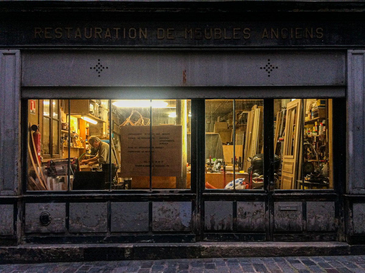 Boutique de restauration de meubles anciens, vitrine éclairée, intérieur encombré.