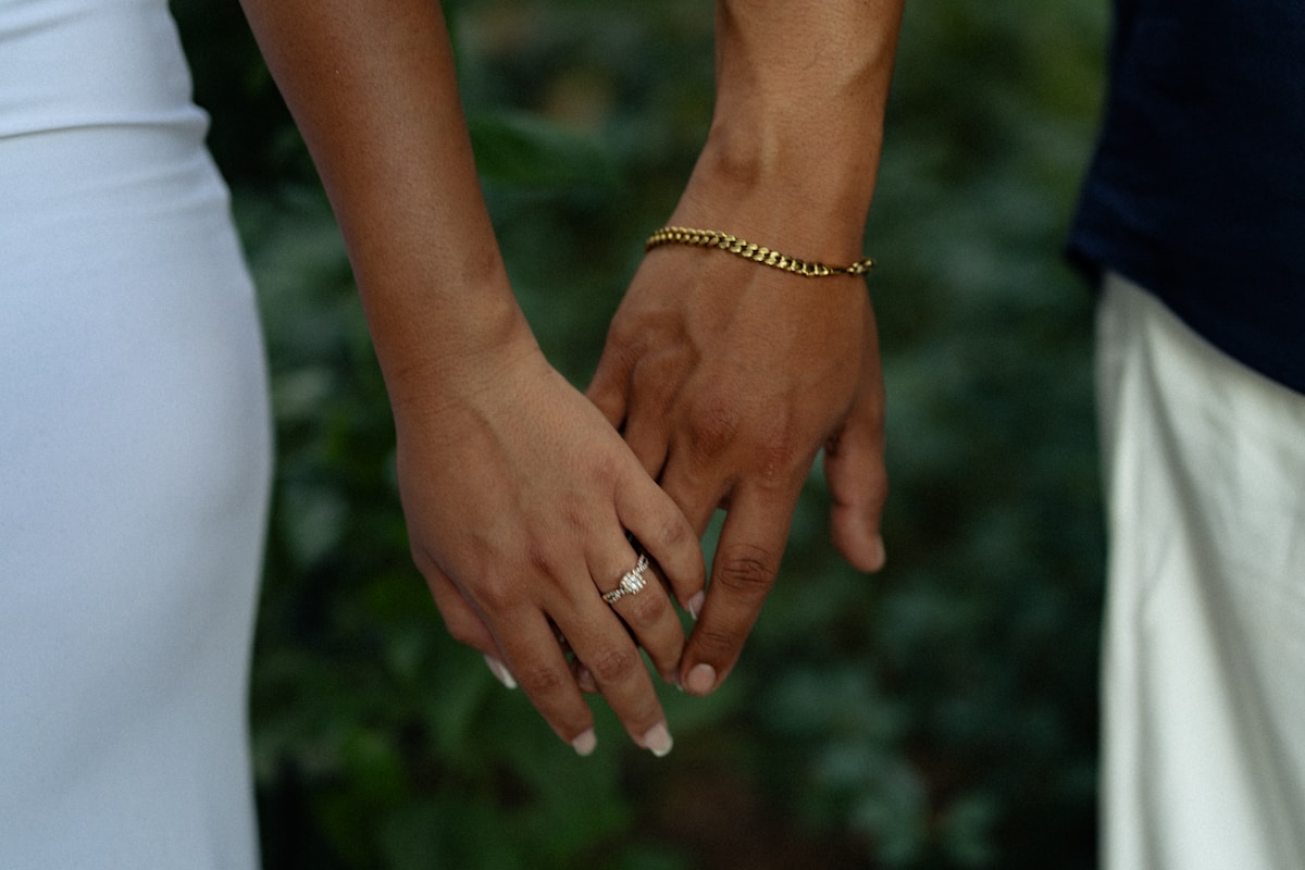 Mains jointes, bague de fiançailles, bracelet doré, décor verdoyant.